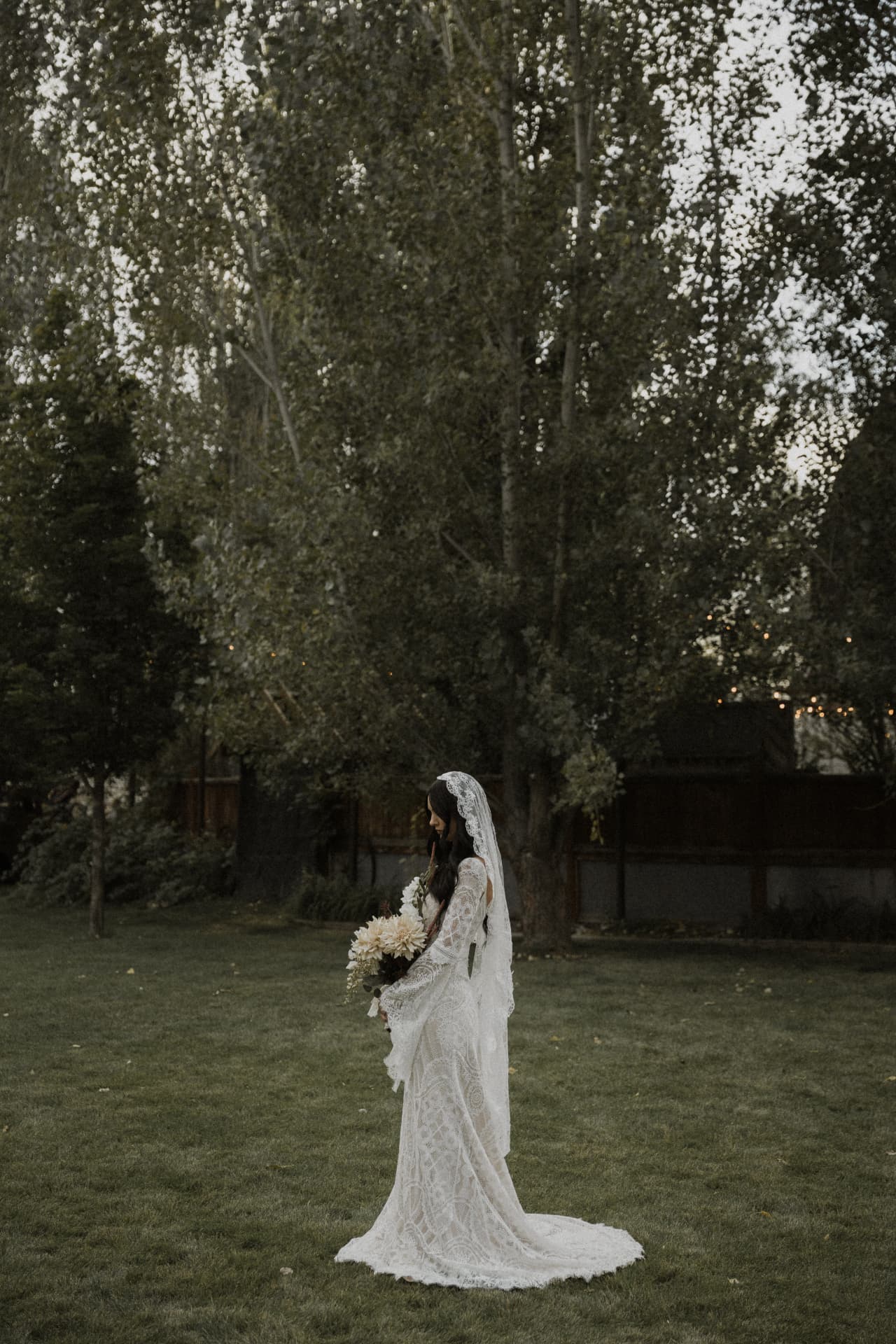 A bride with a long veil standing on a lawn beneath the trees