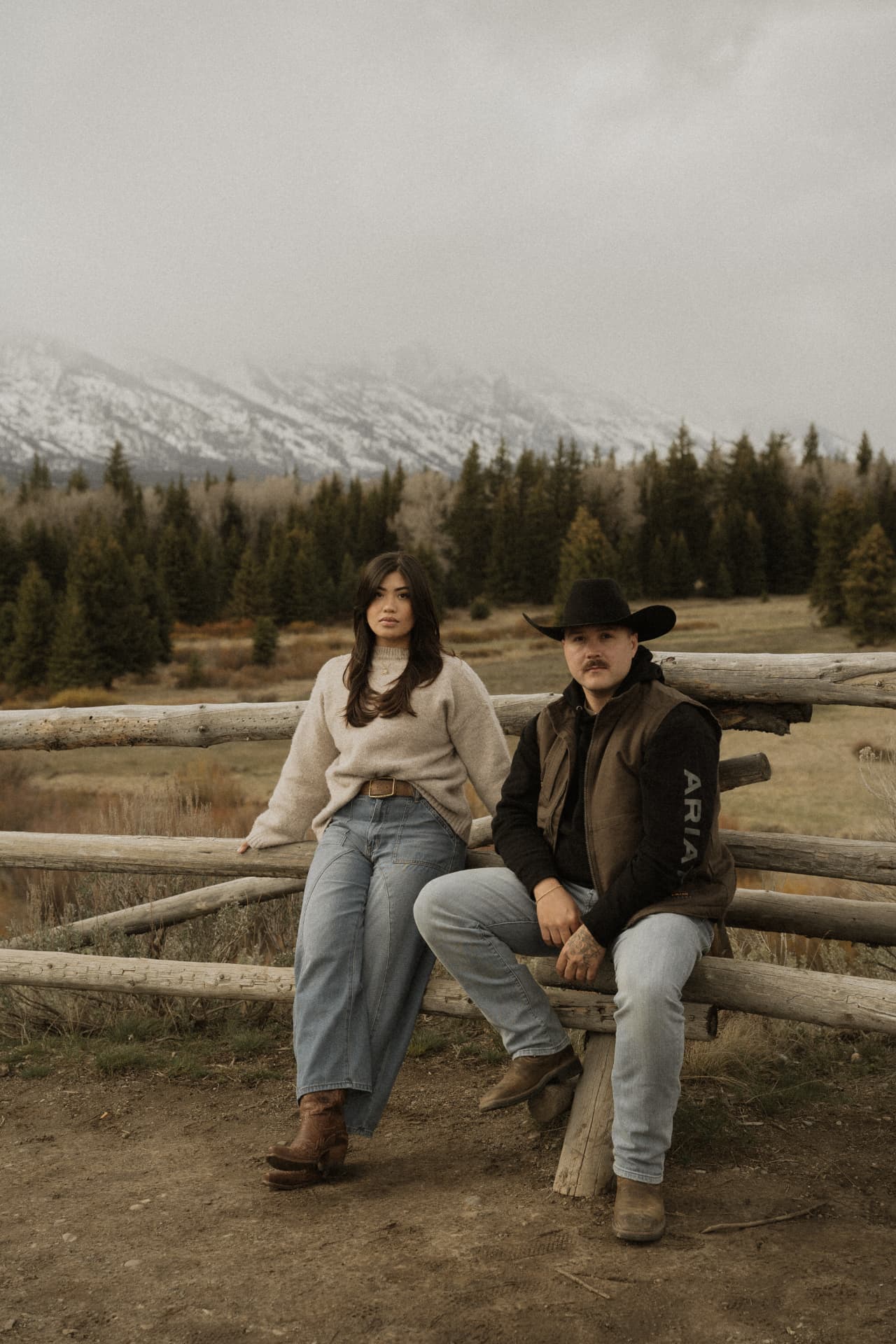 A couple sitting on a wooden fence beneath snowy Teton peaks