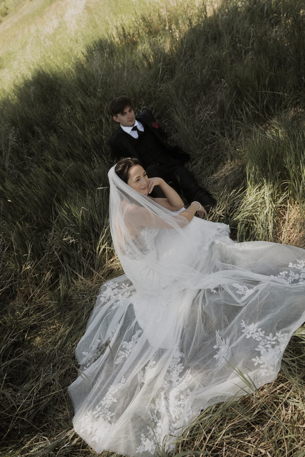 A bride and groom seated in grass with her veil spread around them