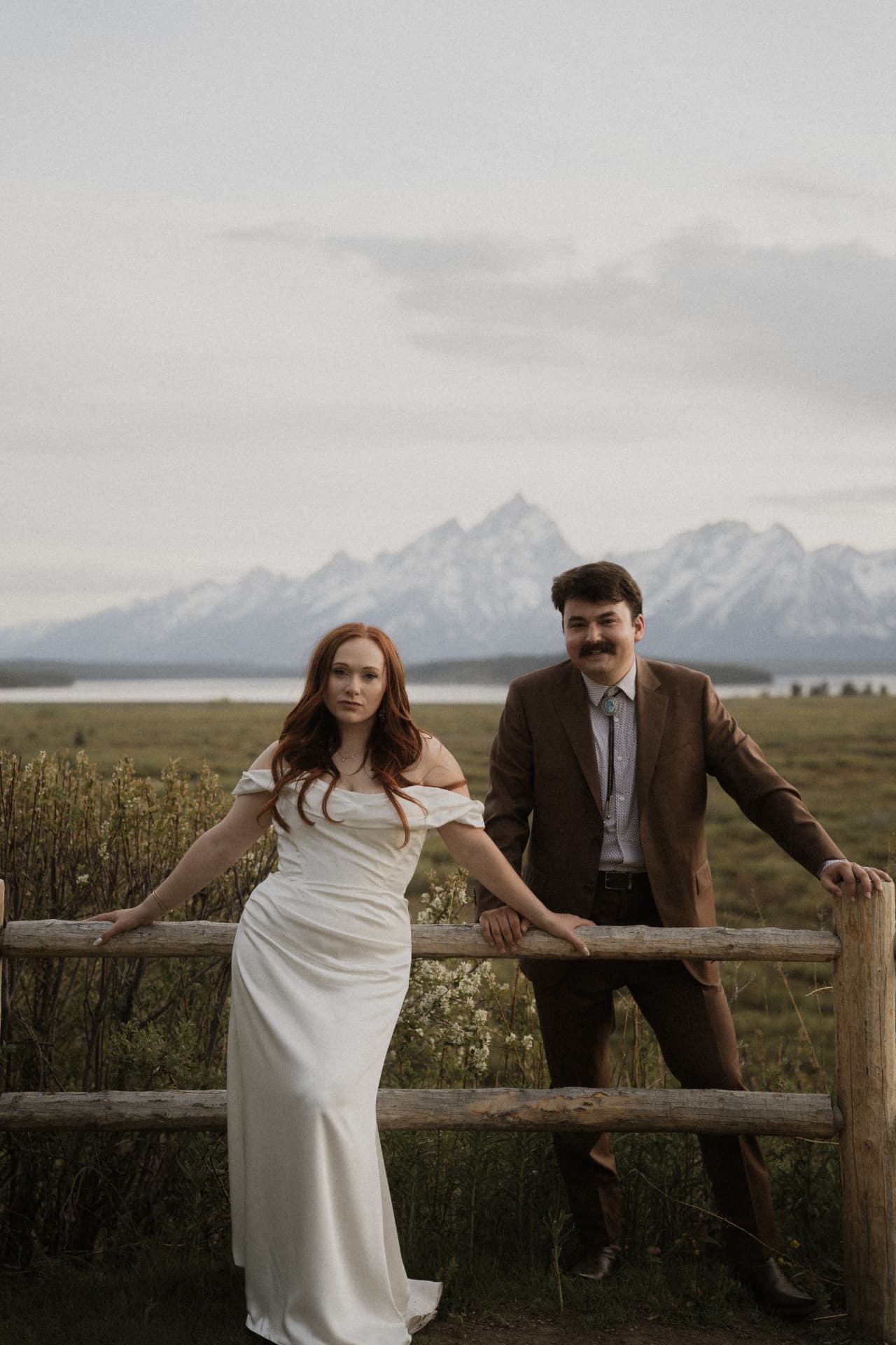 A couple leaning on a wooden fence with the Grand Tetons behind them