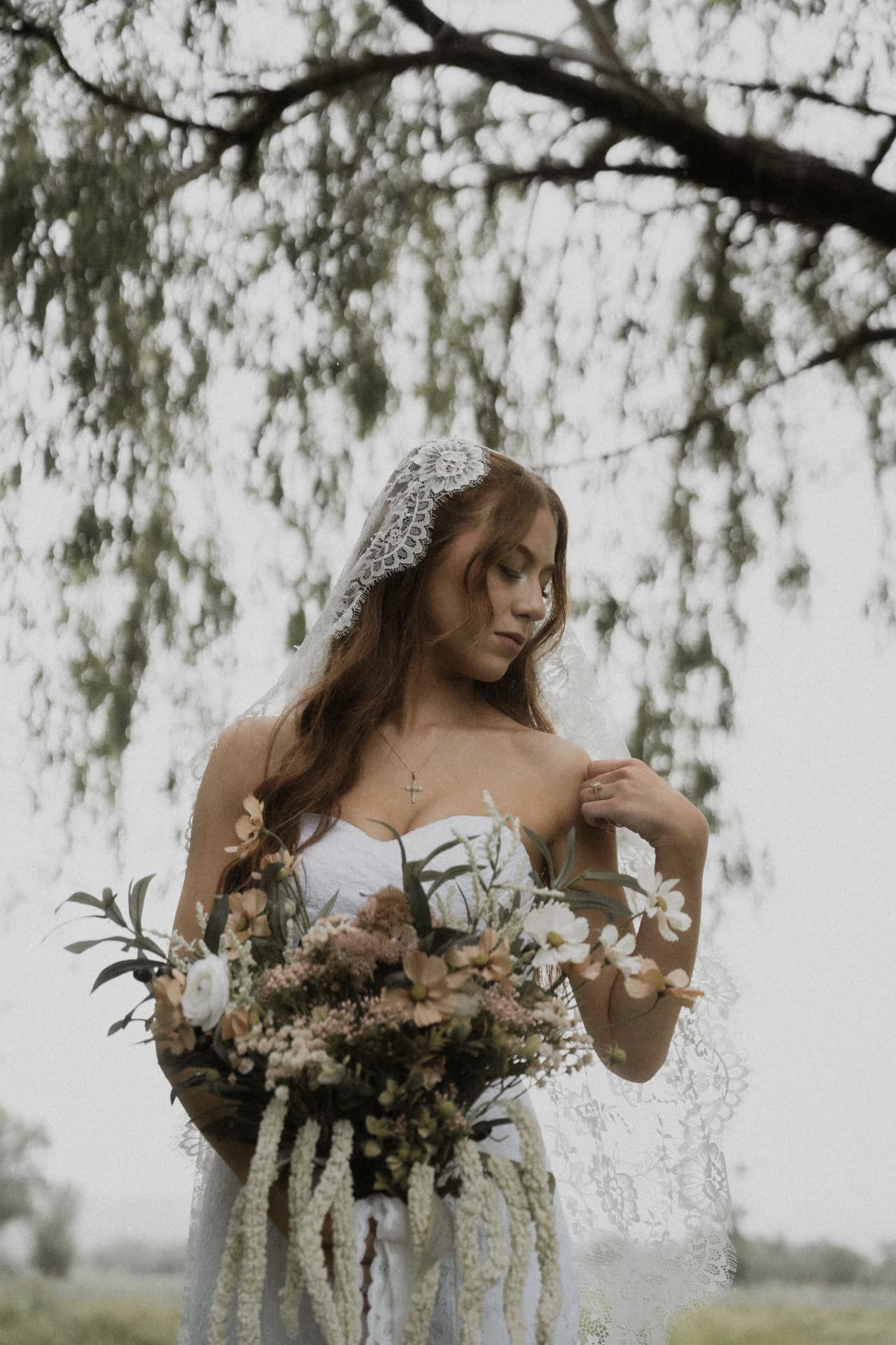 A bride with a lace veil holding a wildflower bouquet beneath a willow tree