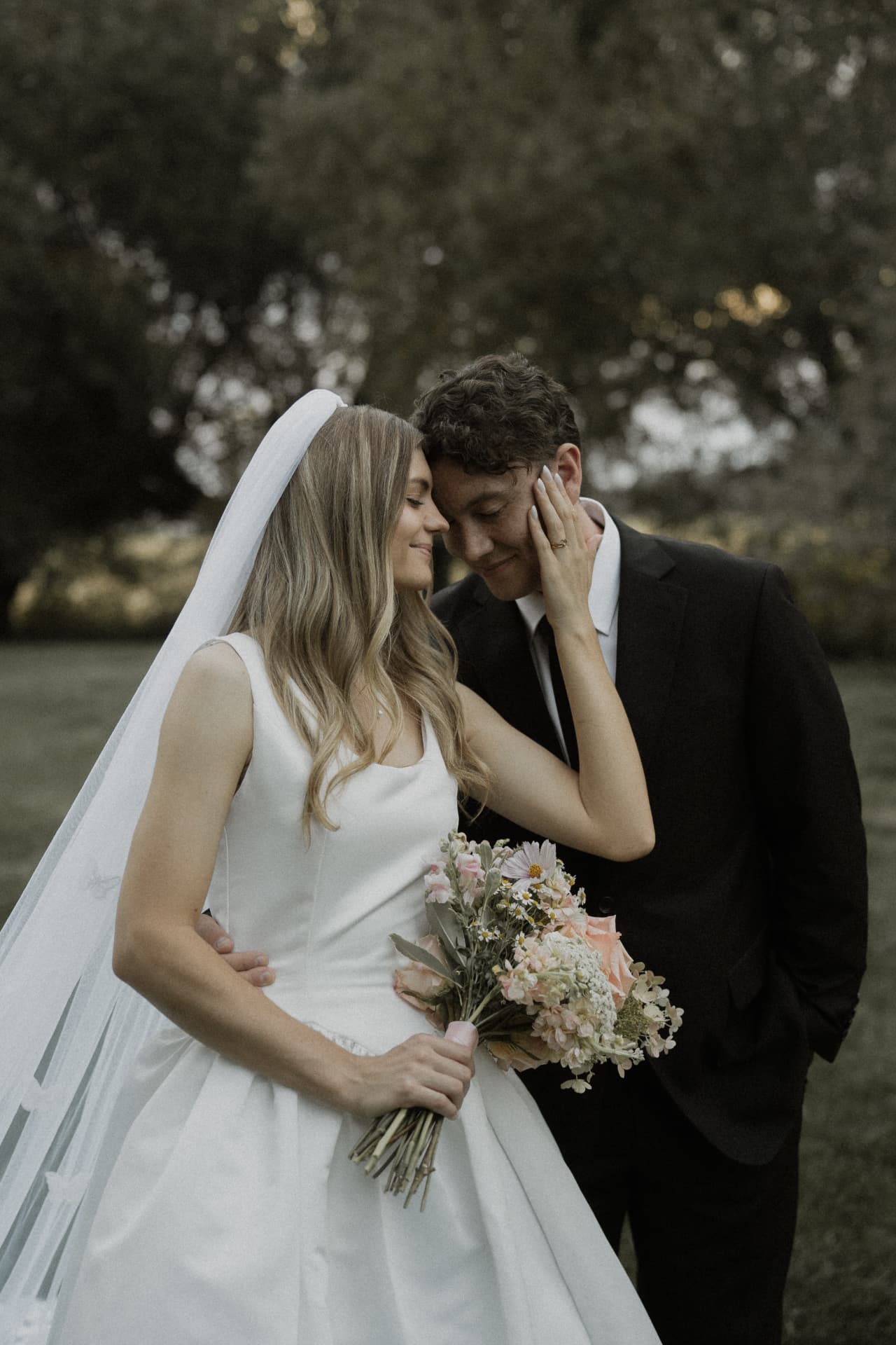 A bride holding flowers and touching the groom’s face beneath the trees